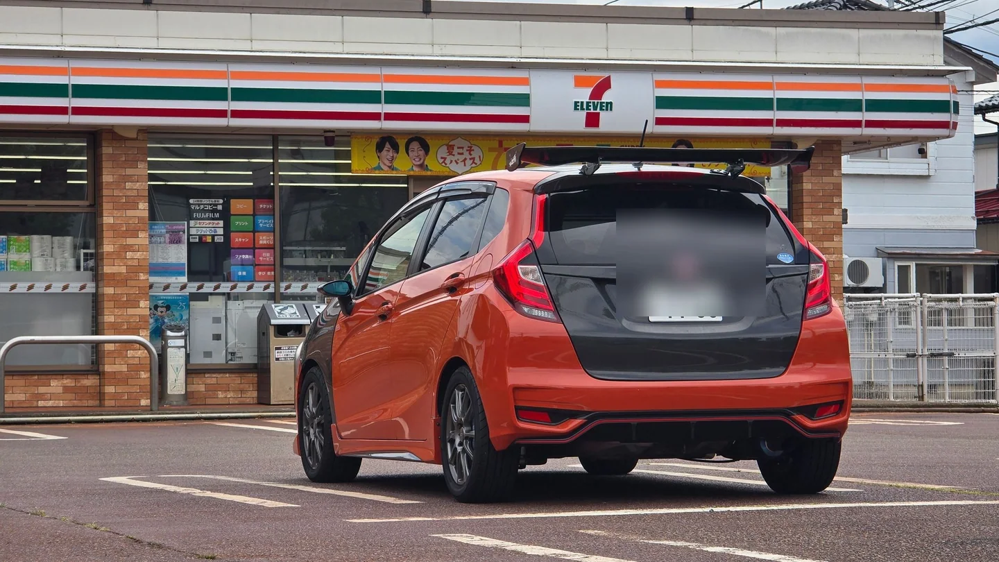 カーボントランクとGTウイングを装着したオレンジのHonda Fitの後ろ姿。ナンバーはぼかされている。