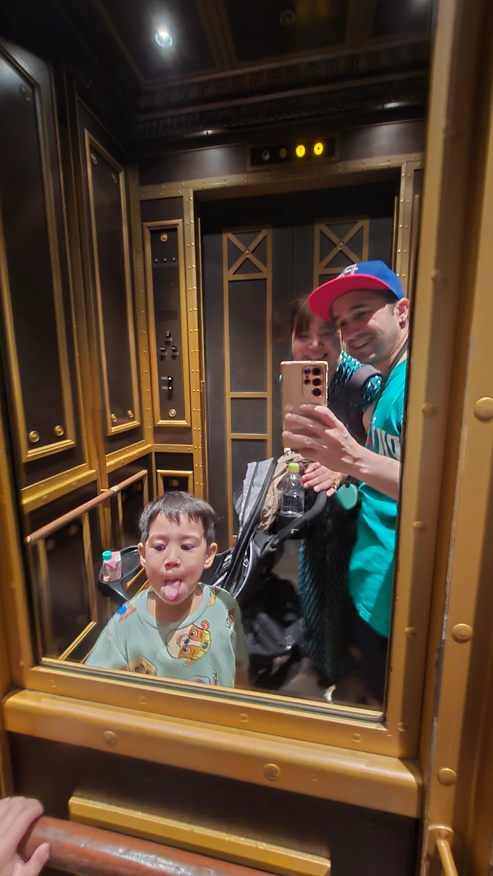 A family takes a playful mirror selfie inside an ornate elevator while the youngest child sticks out his tongue.