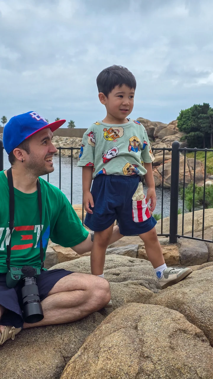 A father smiles up at his youngest son standing proudly on rocks beside the water at Tokyo DisneySea.