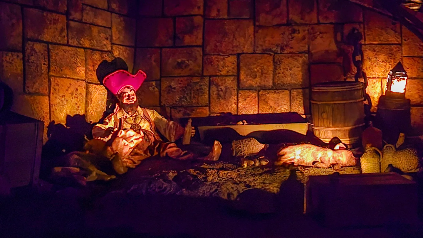 A warmly lit pirate animatronic reclines beside barrels and sacks inside Pirates of the Caribbean.