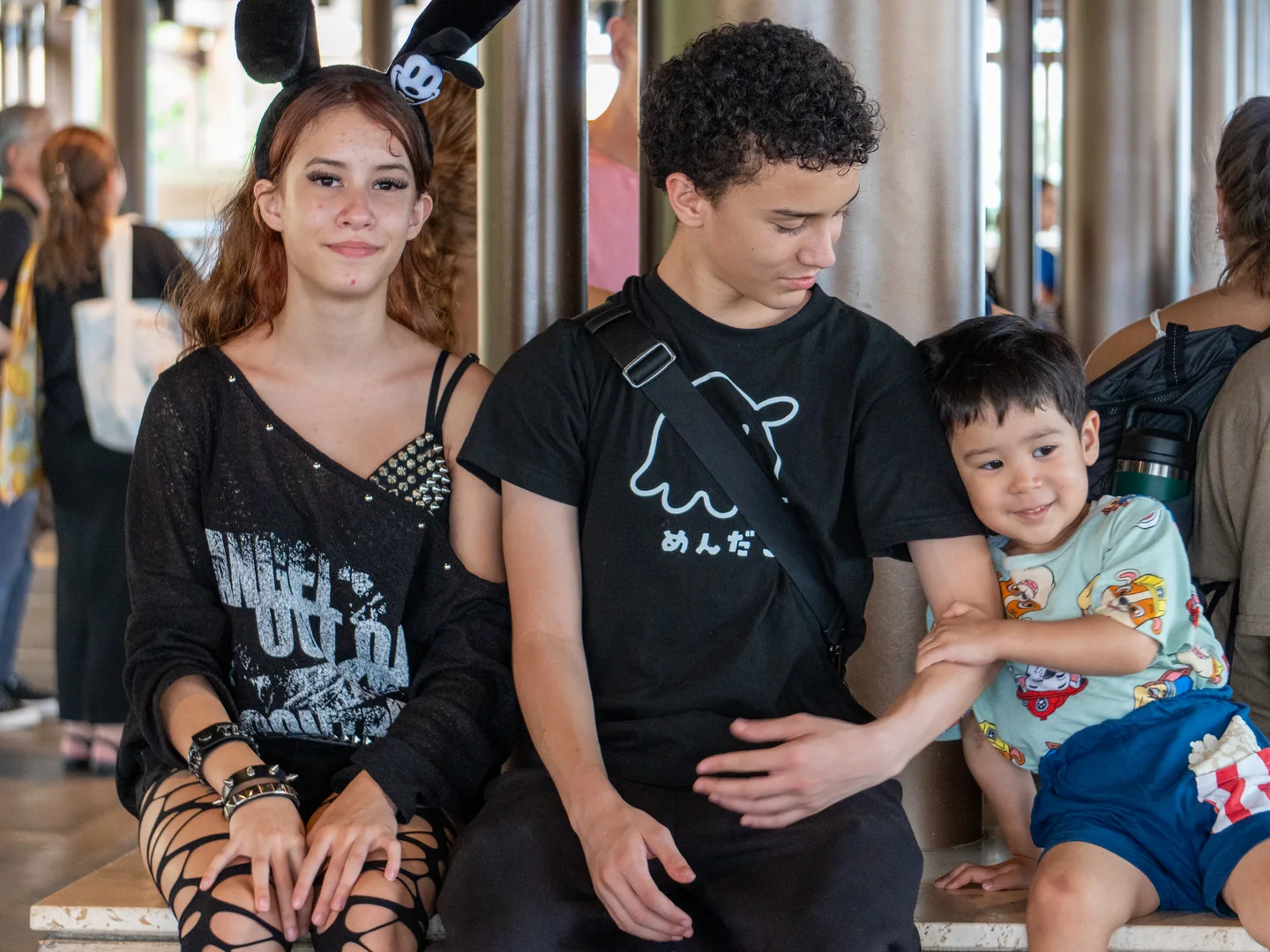 Three siblings sit together during a break, with the youngest holding tightly to his older brother’s arm.